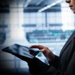 Businesswoman in a dark blazer using a tablet in a modern office; blurred background shows screens and daylight.