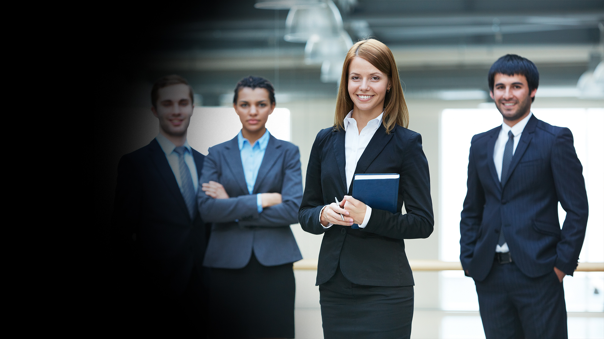 Professional woman in a black suit smiles at the camera, holding a blue notebook and a pen, with three colleagues in the background.
