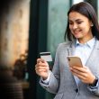 Businesswoman in a gray blazer smiling outdoors while holding a smartphone and a credit card, ready to pay.